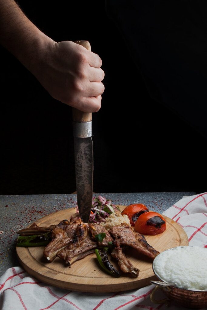 Mão segurando faca em cima da carne de cordeiro na tábua de madeira.