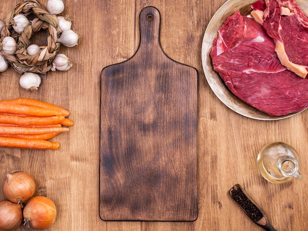 A Importância do Corte Certo na Preparação de Carnes 1 Vista De Cima De Uma Tábua De Corte De Madeira Vintage Com Dois Pedaços De Carne Vermelha, Cercados Por Cenouras Laranjas, Pimentões E Vagens Em Uma Mesa De Madeira.